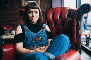 A landscape picture of Ellie Holland sat in a deep red Chesterfield armchair