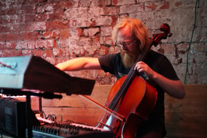 Drew Morgan in front of an exposed red brick wall, playing cello with several synthesisers in shot
