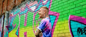 Ben Cross (Crossy) standing side on and smiling with a graffiti'd wall in the background